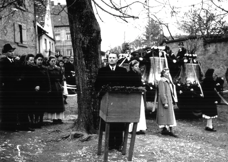 Bürgermeister Kurt Link spricht am Holzpult vor den geschmückten Glocken auf einem Wagen, umgeben von Mädchen und Zuschauern.