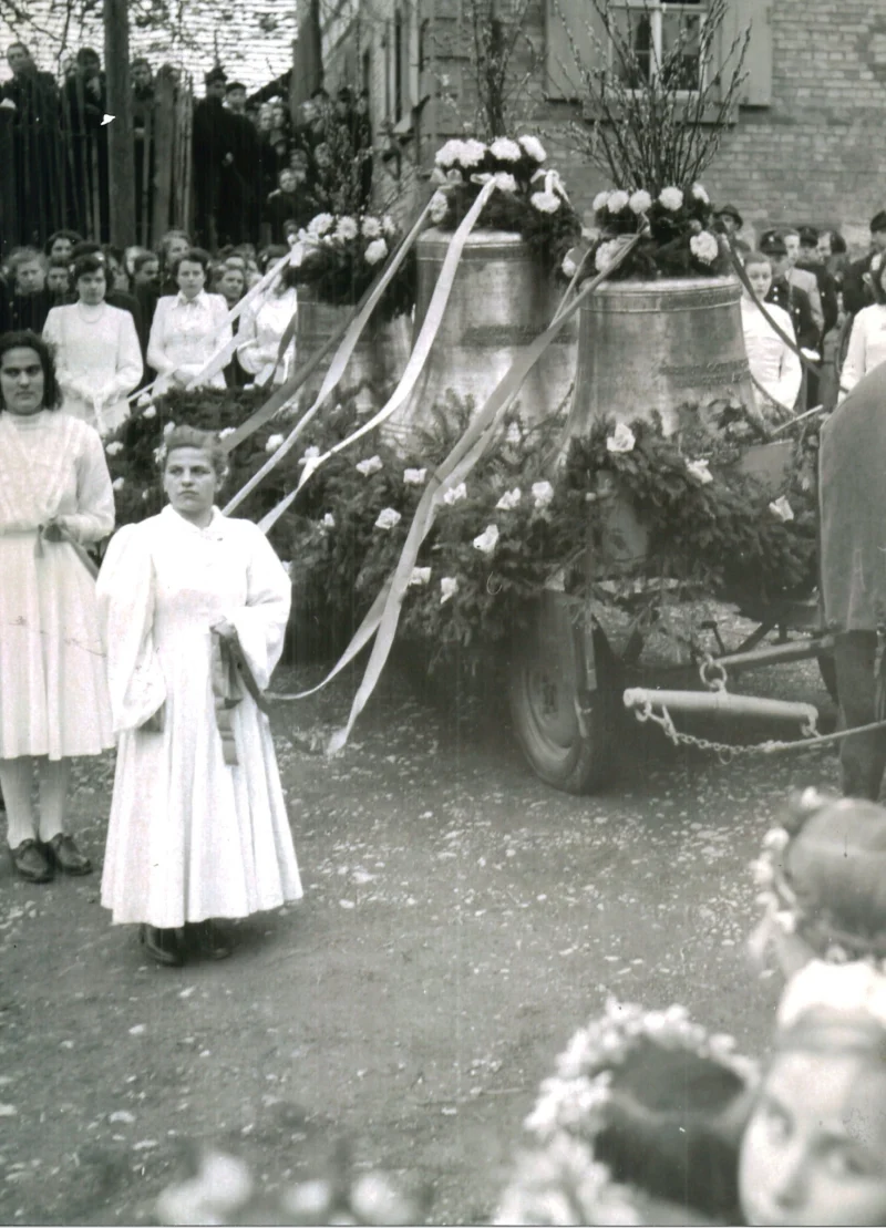 Mädchen in weißen Kleidern mit Blumenkränzen stehen neben den geschmückten Glocken auf einem Wagen.
