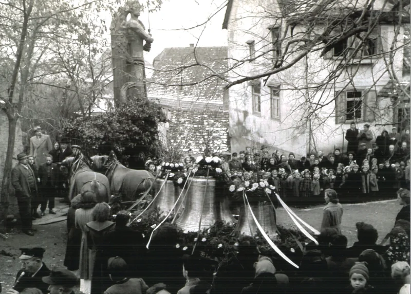 Pferde ziehen den Wagen mit den geschmückten Glocken an einem Denkmal und an Häusern vorbei, ringsum stehen Zuschauer.