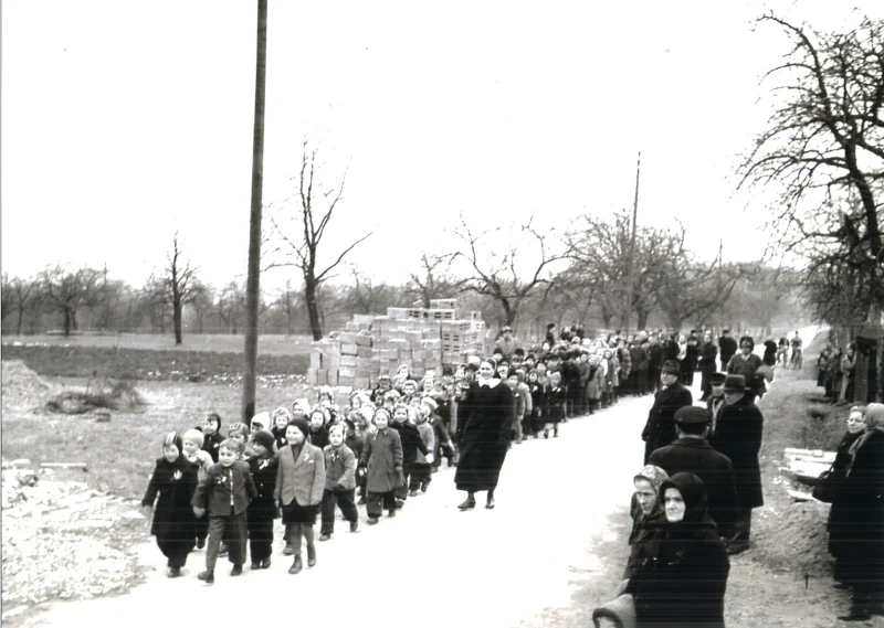 Ein langer Zug von Kindern geht eine Landstraße außerhalb des Orts entlang, am Rand stehen Erwachsene.