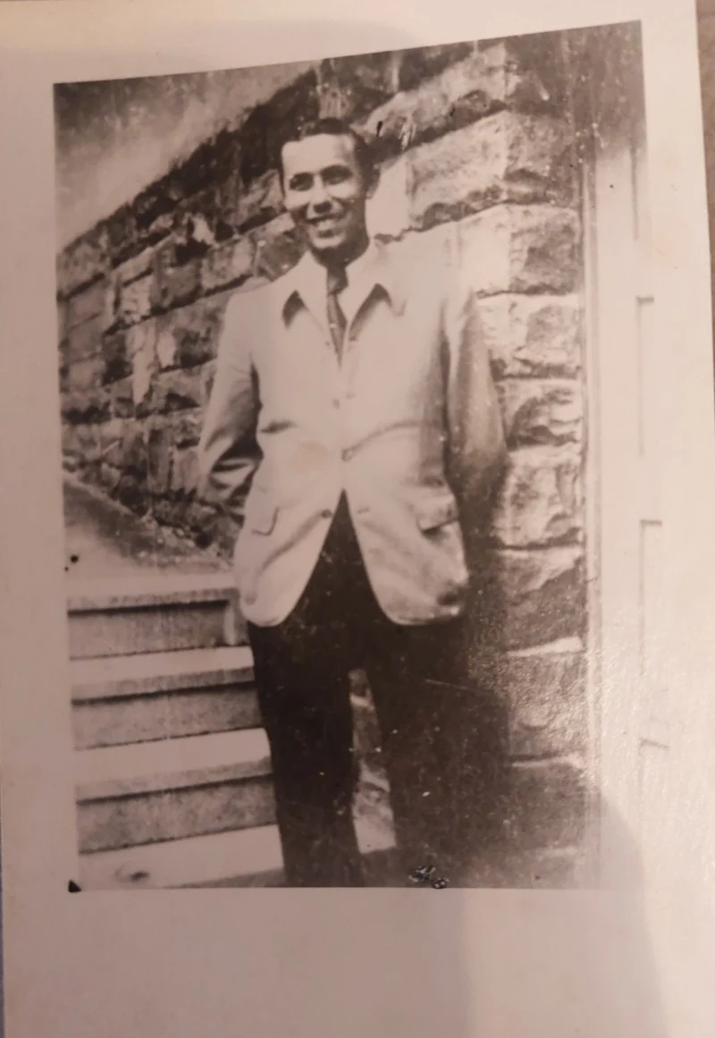 Schwarzweißfoto eines lächelnden jungen Mannes in hellem Sakko mit Krawatte vor einer Bruchsteinmauer neben einer Treppe.