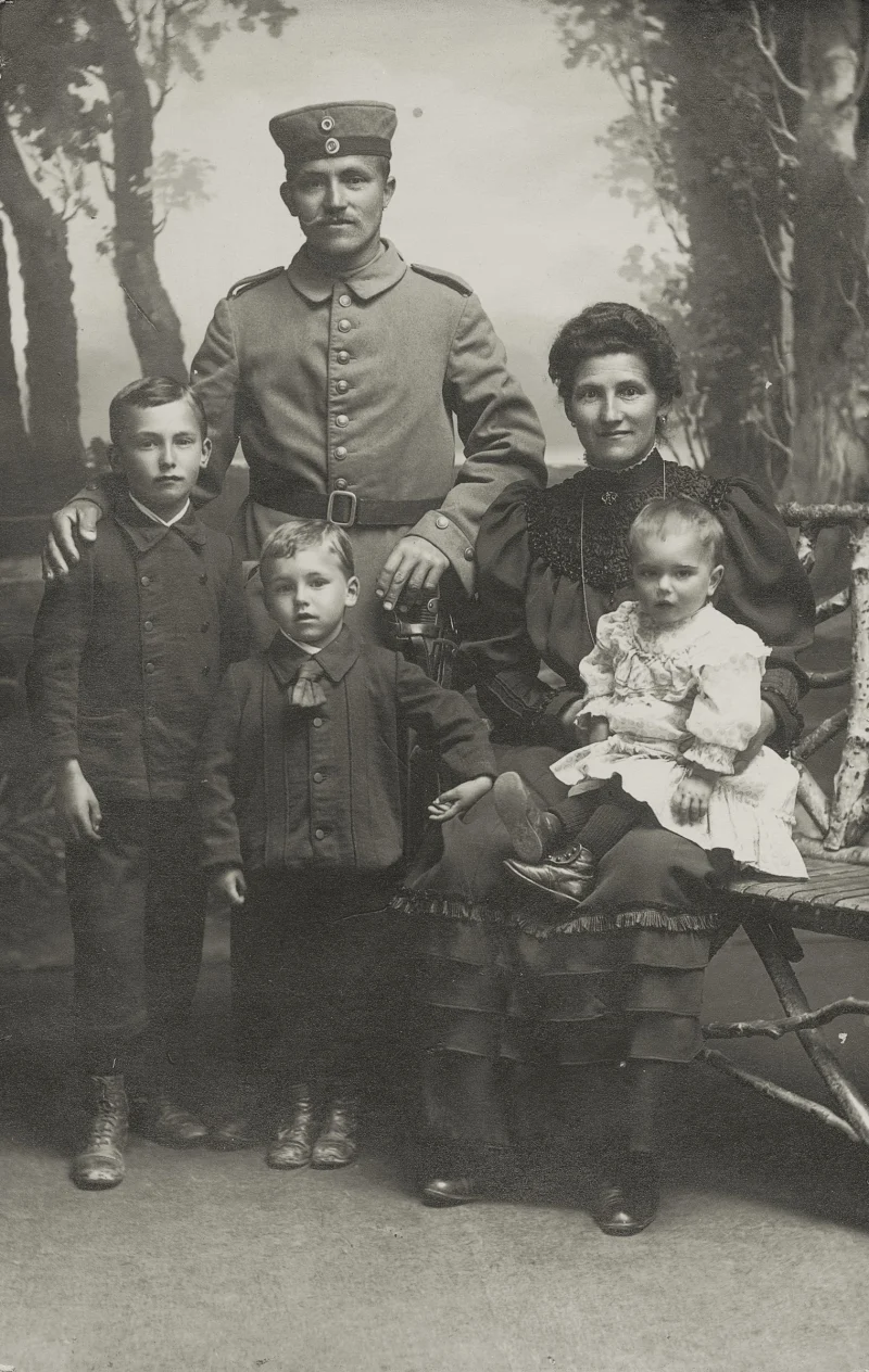 Atelieraufnahme einer Familie: ein Mann in Uniform mit Mütze, daneben eine Frau mit Kleinkind auf dem Schoß, davor zwei Jungen.