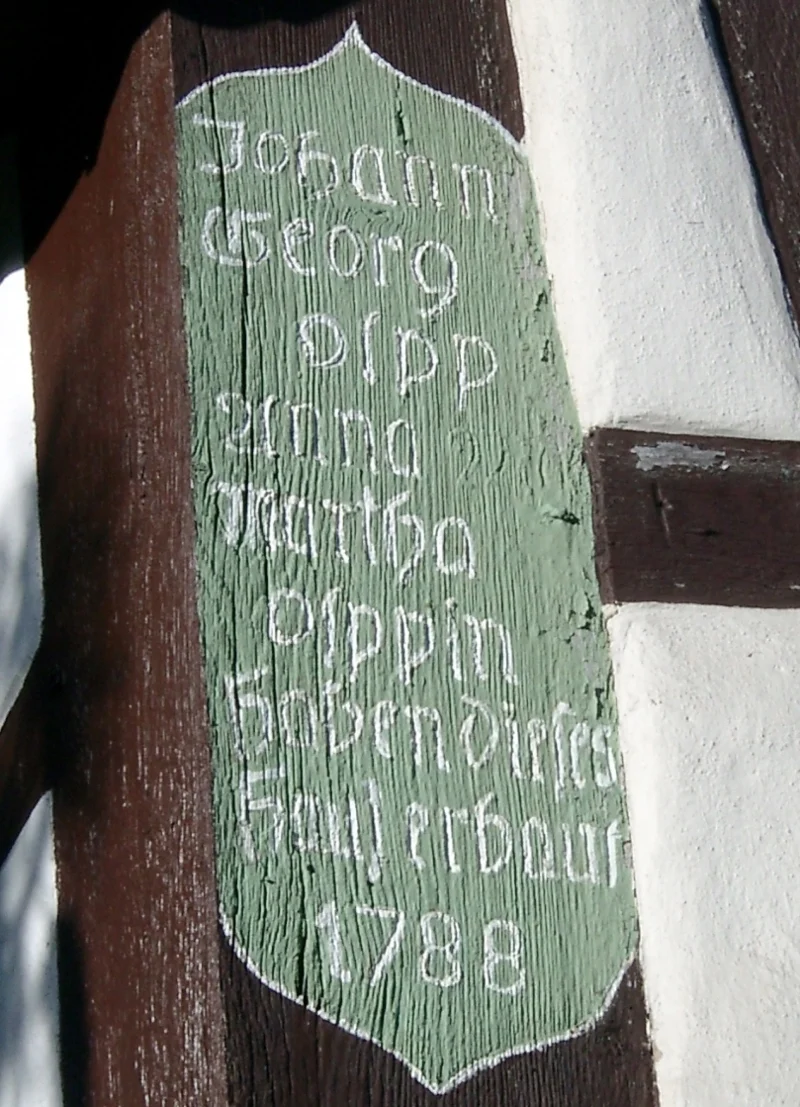 Grün bemaltes Holzschild am Fachwerk: „Johann Georg Olpp, Anna Martha Olppin haben dieses Haus erbaut 1788“.