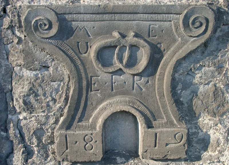 Steinernes Hauszeichen mit Voluten und einer Brezel in der Mitte, darum Initialen und die Jahreszahl 1819.