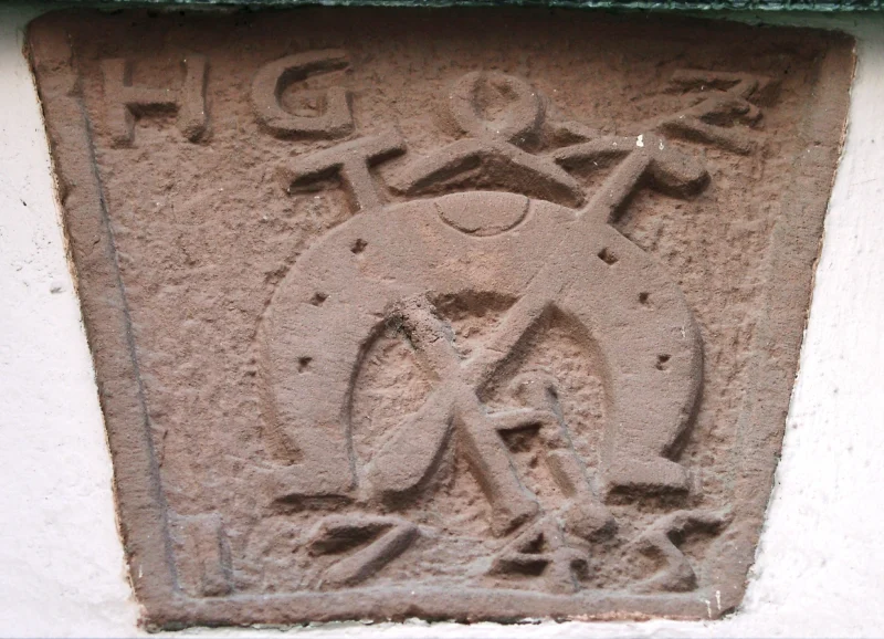 Rotes Sandsteinzeichen mit den Initialen H G Z, einem Hufeisen und gekreuzten Werkzeugen.