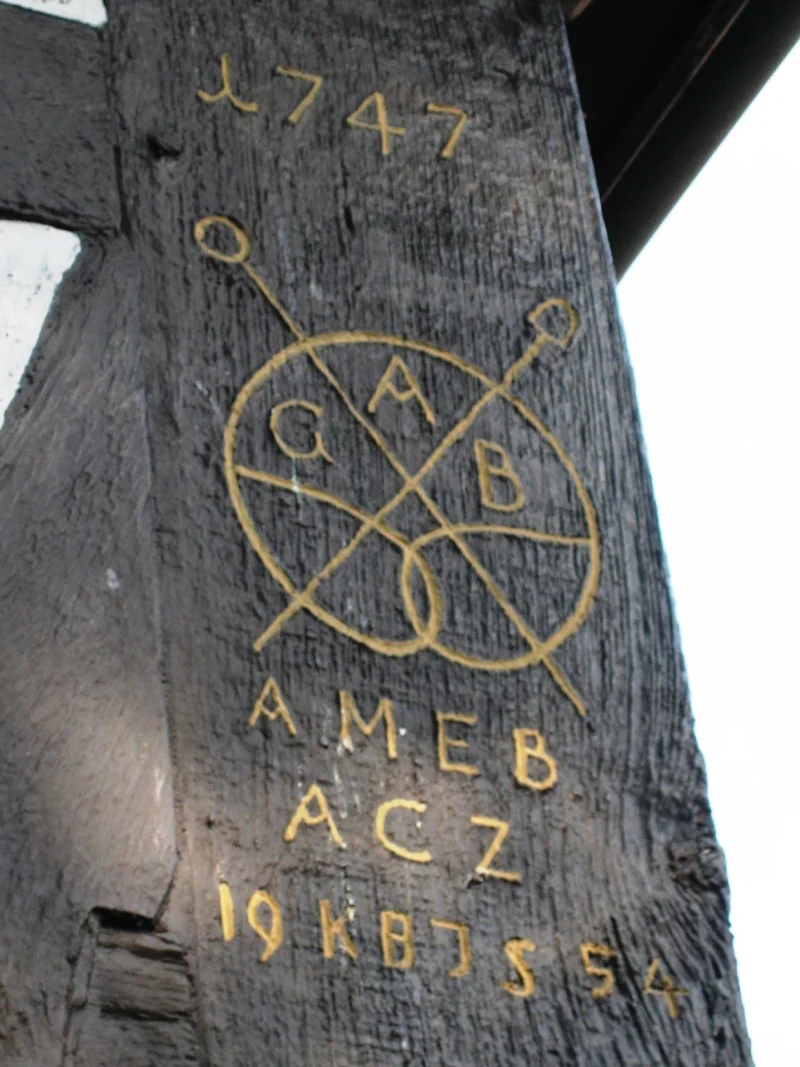 Dunkler Holzbalken mit gelb nachgezogener Inschrift: Jahreszahl 1747, ein Kreis mit gekreuzten Werkzeugen und mehrere Initialen.