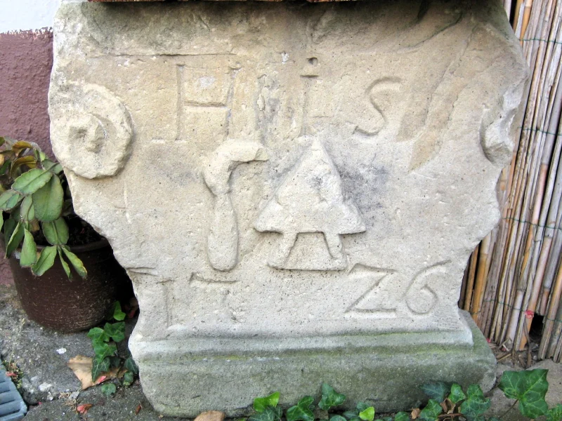 Verwitterte Steinplatte mit Voluten, den Initialen H S und eingemeißelten Werkzeugen.