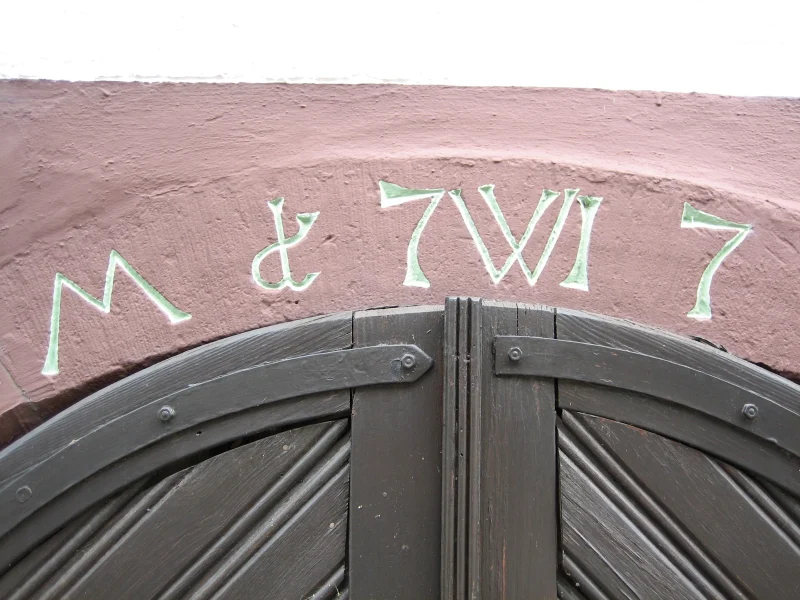 Rot gestrichener Torbogen mit hell nachgezogenen Buchstaben und Ziffern über einem Holztor.