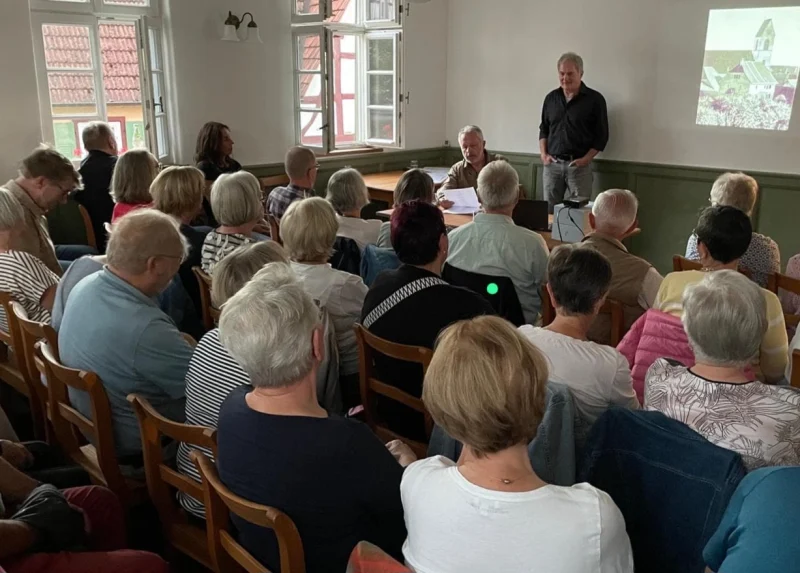 Zuhörerinnen und Zuhörer im Grünen Salon des Kronenschulhauses; vorne liest ein Mann am Tisch, ein zweiter steht neben der Projektionsfläche.