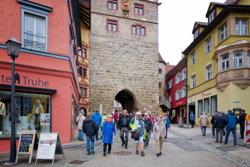 Die Reisegruppe geht durch die Gasse vor dem Schwarzen Tor, einem mittelalterlichen Torturm aus Bruchstein mit Uhr, zwischen farbigen Bürgerhäusern.
